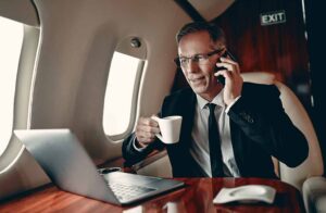 Businessman in a suit sitting inside a private jet, talking on the phone and holding a coffee mug while working on a laptop.