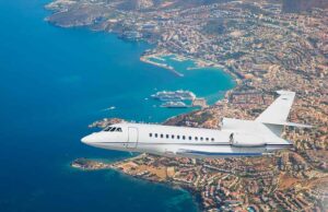Private jet flying over a scenic coastline and city with blue water, urban buildings, and a marina below.
