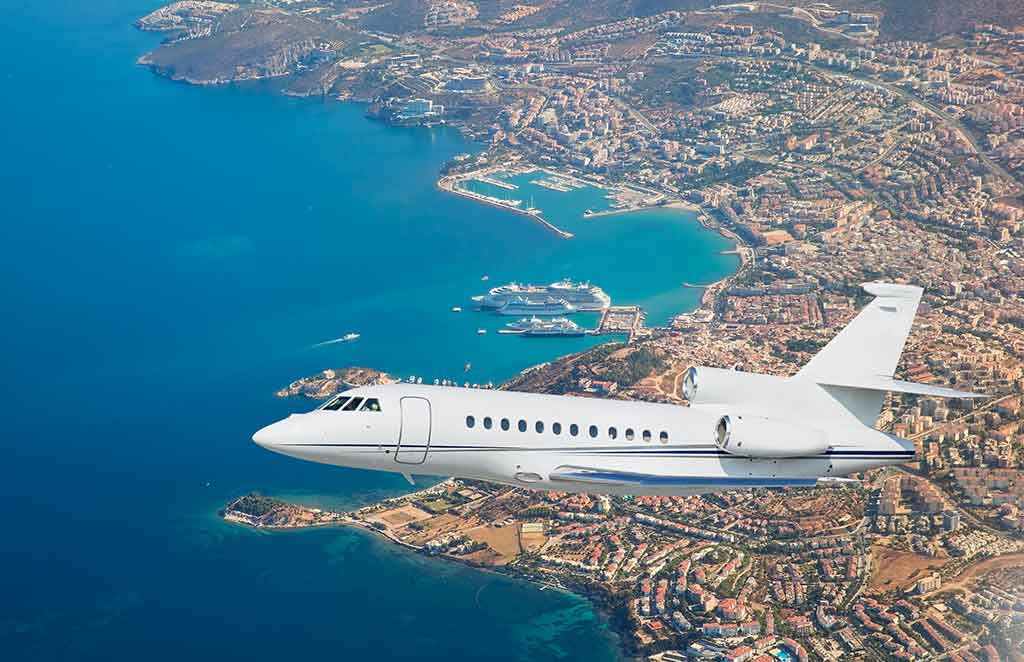 Private jet flying over a scenic coastline and city with blue water, urban buildings, and a marina below.