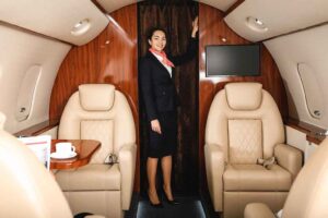 Smiling flight attendant standing inside a private jet cabin, next to two plush leather seats and a wood-paneled interior.