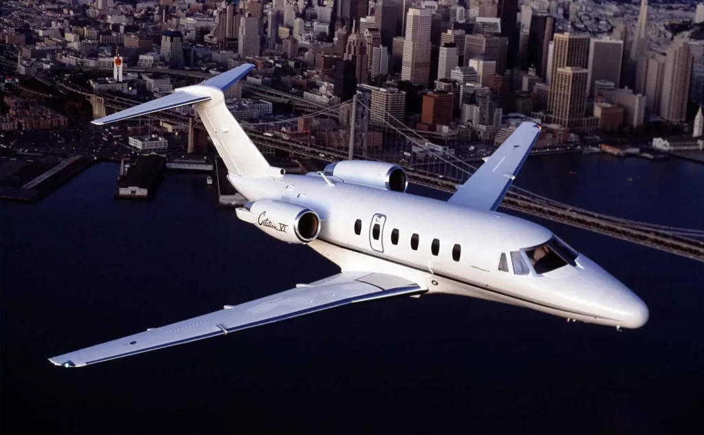 White private jet flying above a cityscape with a prominent bridge and skyscrapers in the background